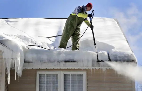 A man atop a house roof uses a steam wand to remove ice dams.